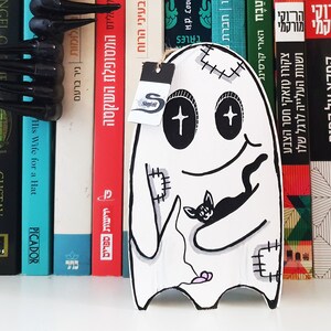 Puede incluir: Un fantasma blanco con ojos negros y una boca cosida sosteniendo un gato negro. El fantasma tiene un coraz&oacute;n rosa y un dise&ntilde;o cosido en su cuerpo. El fantasma est&aacute; en una estanter&iacute;a con libros detr&aacute;s.