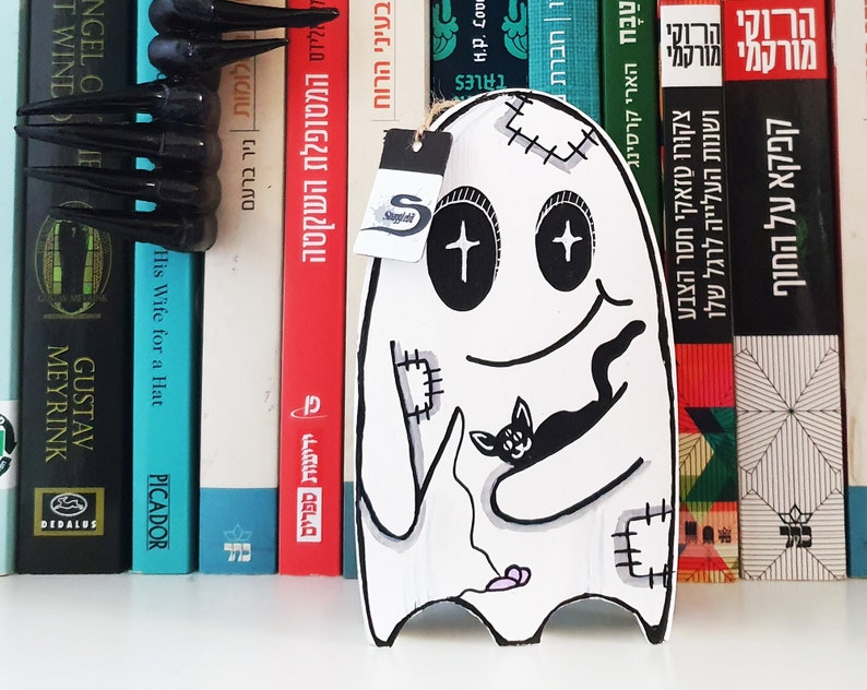 Puede incluir: Un fantasma blanco con ojos negros y una boca cosida sosteniendo un gato negro. El fantasma tiene un coraz&oacute;n rosa y un dise&ntilde;o cosido en su cuerpo. El fantasma est&aacute; en una estanter&iacute;a con libros detr&aacute;s.