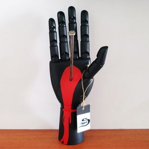 May include: A black wooden hand sculpture with a red painted design and a nail through the palm. A small tag is attached to the wrist. The sculpture is displayed on a wooden surface against a white background.