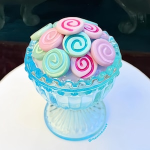 May include: A close-up of a light blue glass bowl filled with colorful, swirled candies. The candies are pink, blue, and green. The bowl sits on a white surface. Two other bowls filled with candies are in the background.