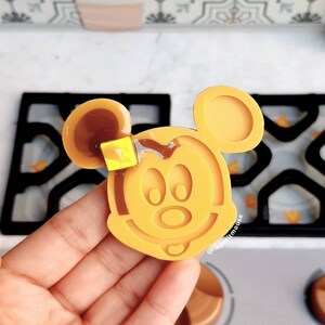May include: A yellow silicone mold shaped like Mickey Mouse's head. The mold is on a white surface with a black waffle iron in the background.