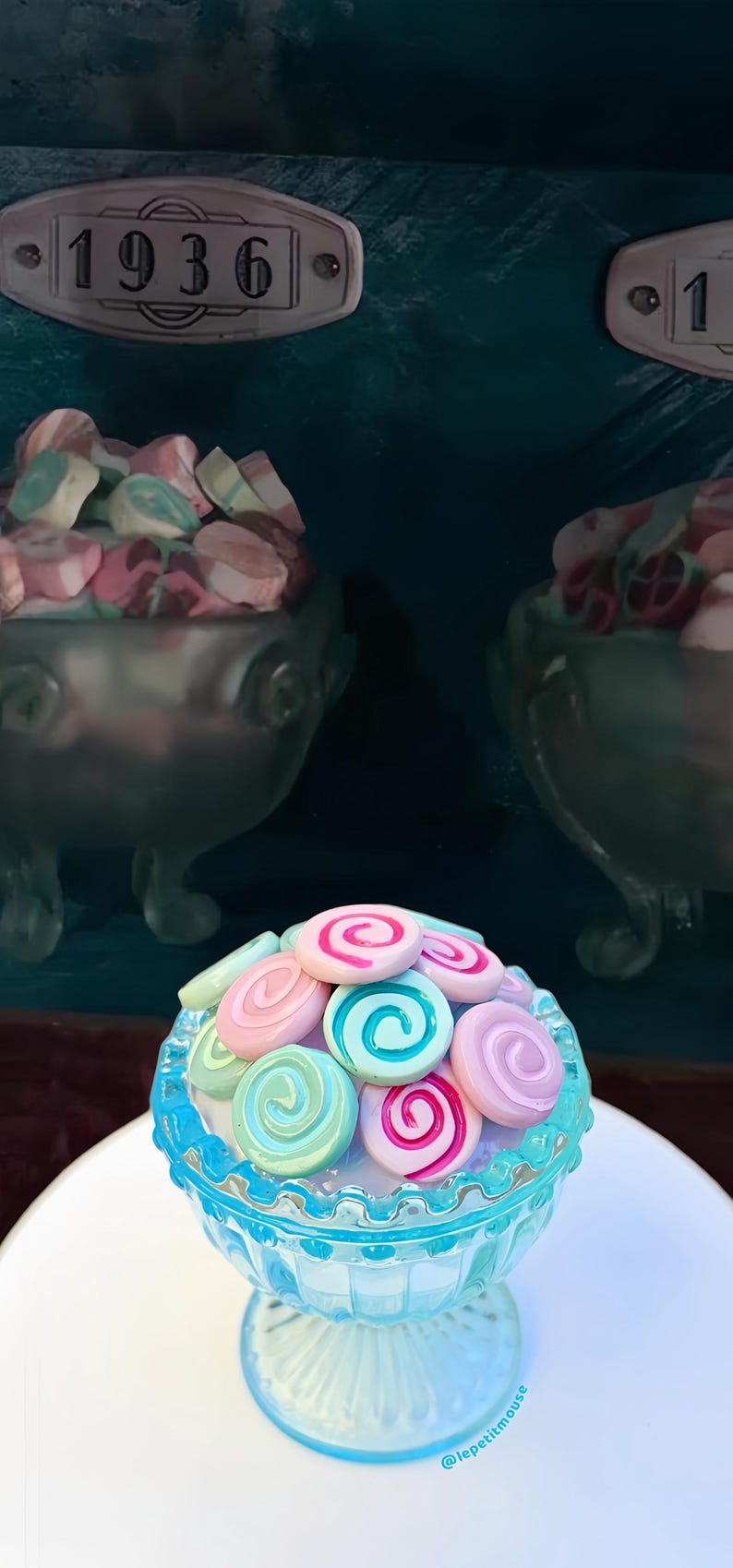 May include: A close-up of a light blue glass bowl filled with colorful, swirled candies. The candies are pink, blue, and green. The bowl sits on a white surface. Two other bowls filled with candies are in the background.