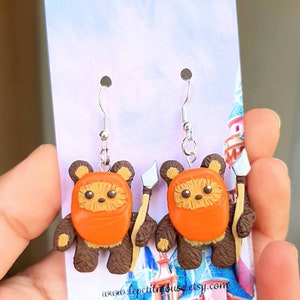 May include: A pair of dangle earrings featuring a brown and orange Ewok character from Star Wars. Each Ewok is holding a small, silver-colored weapon. The earrings are on a white card with a pink and blue background.