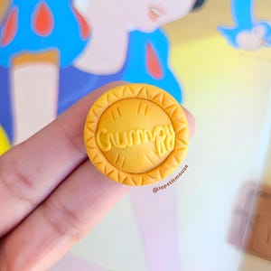 May include: A round, yellow charm with the word "Grumpy" in a stylized font. The charm is held in front of a Snow White illustration. The charm has a textured border and is likely a collectible or accessory.