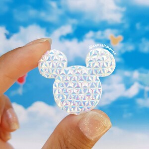 May include: A white iridescent Mickey Mouse head shaped pin with a geometric pattern. The pin is being held by a hand against a blue sky background.