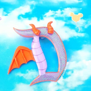 May include: A purple and orange glittery letter "D" shaped pin, featuring a dragon design. The pin has orange horns and wings, with a light purple body. A yellow Mickey Mouse balloon is in the background against a blue sky.