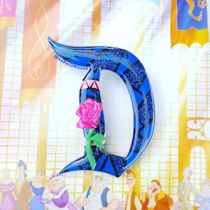 Könnte beinhalten: Ein großer, blauer Buchstabe "D" mit glitzerndem Finish und geometrischem Design, mit einer rosa Rose. Der Hintergrund zeigt eine Cartoon-Illustration von Charakteren und einem Schlossinnenraum, mit dem Text "@LePetitMouse".