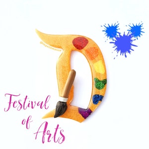 May include: A gold glitter letter D shaped like a paint palette with a paintbrush. The letter D is decorated with colorful glitter and a rainbow paint palette. The text "Festival of Arts" is written below the letter D.