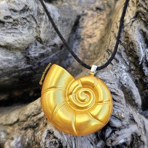 Peut inclure: Un collier pendentif doré avec un motif de coquillage en spirale. Le pendentif est suspendu à un cordon noir. Le fond est une pierre grise texturée.