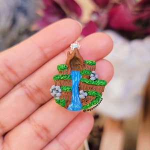 May include: A miniature waterfall scene made of polymer clay. The waterfall is blue and flows down a brown cliff face with green foliage. There are gray rocks on the cliff face. The scene is attached to a silver chain.