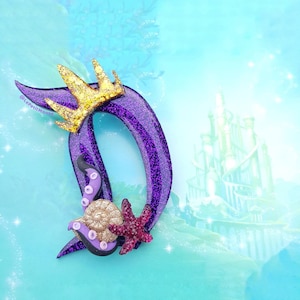 May include: A purple glittery letter "D" with a gold crown, a seashell, a starfish, and tentacles. The background is a light blue with a castle illustration. The letter has a glittery texture.