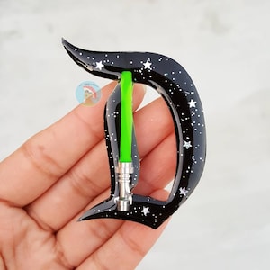 May include: A black letter "D" with a starry night design and white stars. A green lightsaber is in the center of the letter. The letter is approximately 3 inches tall.