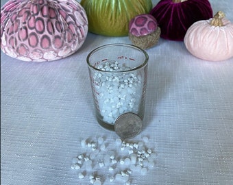 May include: A glass measuring cup filled with white plastic pellets, with a few pellets scattered on a white tablecloth. There are four velvet pumpkins in the background, one pink, one green, one purple, and one with a pink and white pattern.