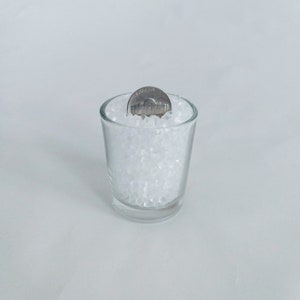 May include: A clear glass shot glass filled with white plastic beads. A silver US penny is partially submerged in the beads.