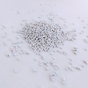 May include: A close-up of a pile of small, white plastic pellets. The pellets are scattered on a white surface.
