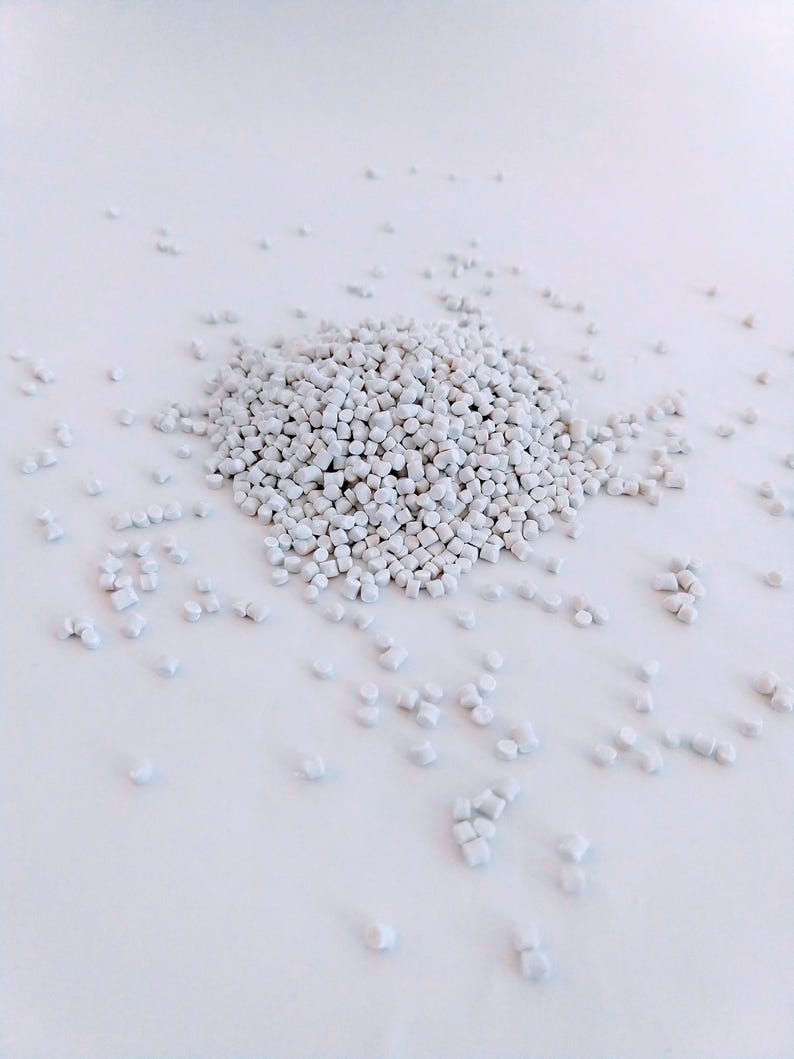 May include: A close-up of a pile of small, white plastic pellets. The pellets are scattered on a white surface.