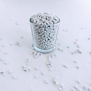 May include: A glass shot glass filled with white plastic pellets. The pellets are scattered on a white surface.