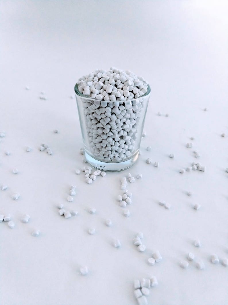 May include: A glass shot glass filled with white plastic pellets. The pellets are scattered on a white surface.
