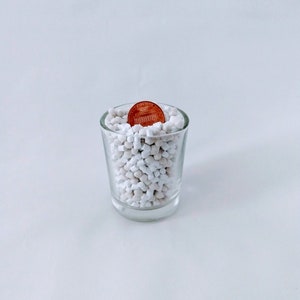 May include: A clear shot glass filled with small white pebbles and a copper penny. The penny is centered in the glass, with the Lincoln Memorial side facing up. The glass sits on a white surface.