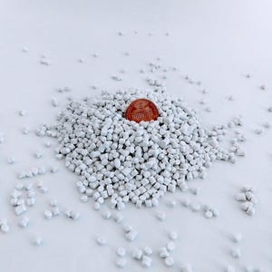 May include: A pile of white plastic pellets with a US penny on top. The pellets are small and round, and they are scattered on a white surface.