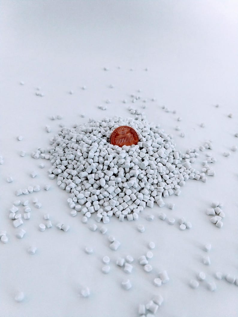 May include: A pile of white plastic pellets with a US penny on top. The pellets are small and round, and they are scattered on a white surface.