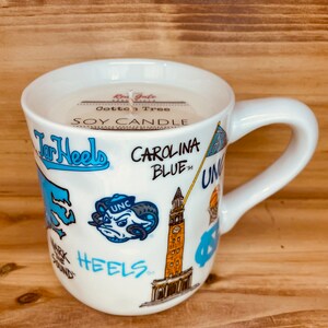 Peut inclure: Une tasse en c&eacute;ramique blanche avec une anse, remplie d'une bougie de soja. La tasse est d&eacute;cor&eacute;e de graphiques bleus et blancs, avec les mots "Tar Heels" et "Carolina Blue". La bougie porte une &eacute;tiquette "Cotton Tree".