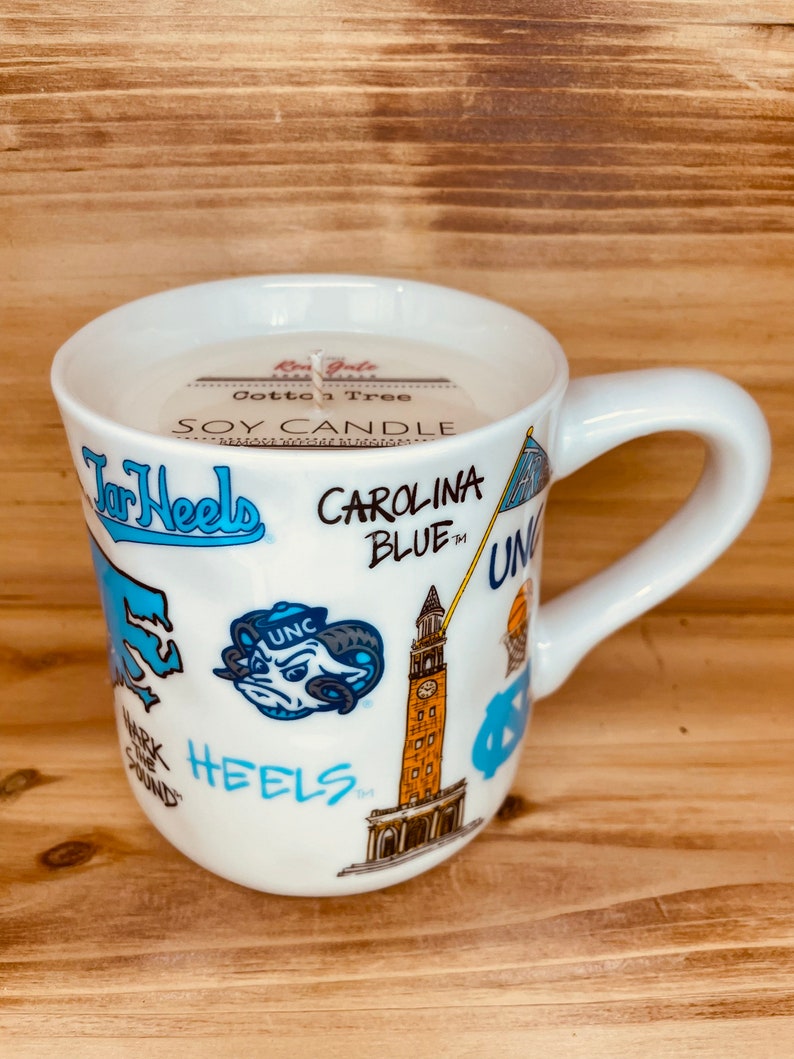 Peut inclure: Une tasse en c&eacute;ramique blanche avec une anse, remplie d'une bougie de soja. La tasse est d&eacute;cor&eacute;e de graphiques bleus et blancs, avec les mots "Tar Heels" et "Carolina Blue". La bougie porte une &eacute;tiquette "Cotton Tree".