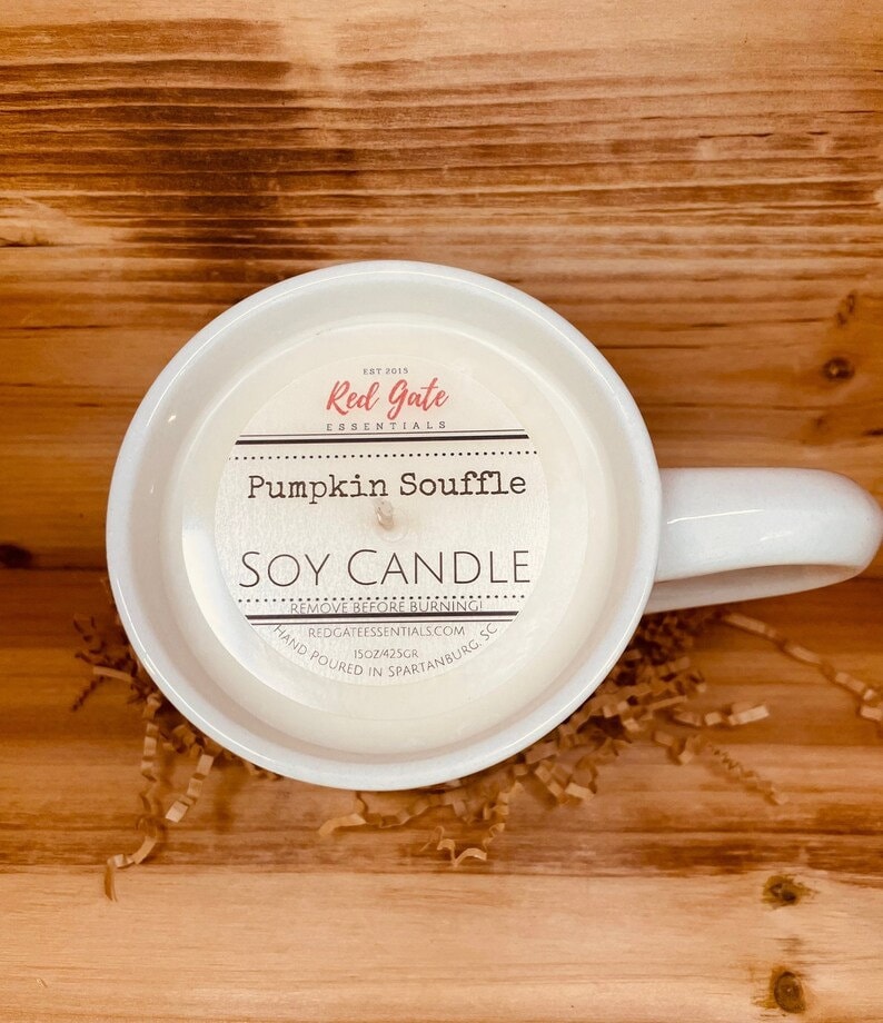 May include: White ceramic mug candle with a label that reads "Red Gate Essentials" and "Pumpkin Souffle Soy Candle". The label also includes the text "Remove Before Burning", "Redgateessentials.com", "1502/425gr", and "Hand Poured in Spartanburg, SC".