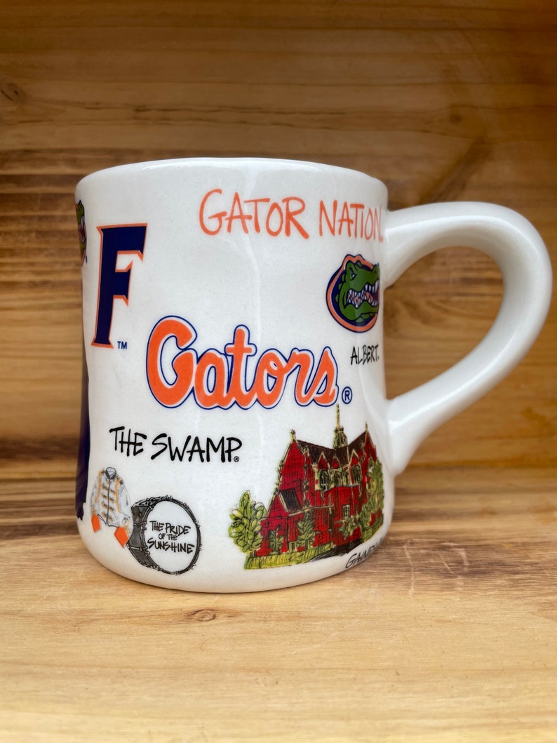 May include: White ceramic mug with a colorful design featuring the words "Gator Nation", "Gators", "The Swamp", and "The Pride of the Sunshine". The mug also features a cartoon alligator, a building, and other details.