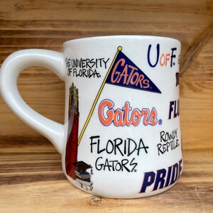 May include: White ceramic mug with a University of Florida Gators theme. The mug features a red and white building, a blue and white flag with the word "Gators" on it, and the words "University of Florida", "Gators", "Florida Gators", "Rowdy Reptile", and "Pride".