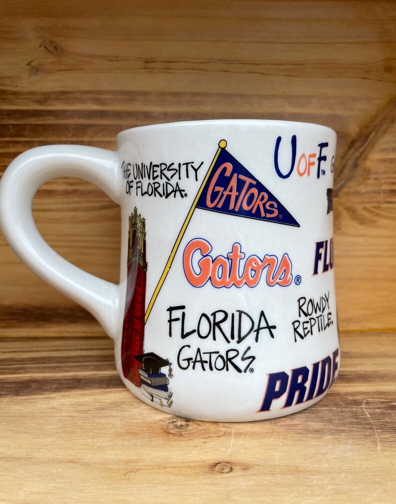 May include: White ceramic mug with a University of Florida Gators theme. The mug features a red and white building, a blue and white flag with the word "Gators" on it, and the words "University of Florida", "Gators", "Florida Gators", "Rowdy Reptile", and "Pride".