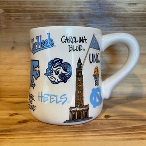 Peut inclure: Mug en c&eacute;ramique blanche orn&eacute; de graphiques et de texte bleu Carolina, dont "Tar Heels", "Carolina Blue" et "UNC". La tasse a une anse incurv&eacute;e et est d&eacute;cor&eacute;e d'illustrations d'un b&eacute;lier, d'un ballon de basket et d'une tour de l'horloge.