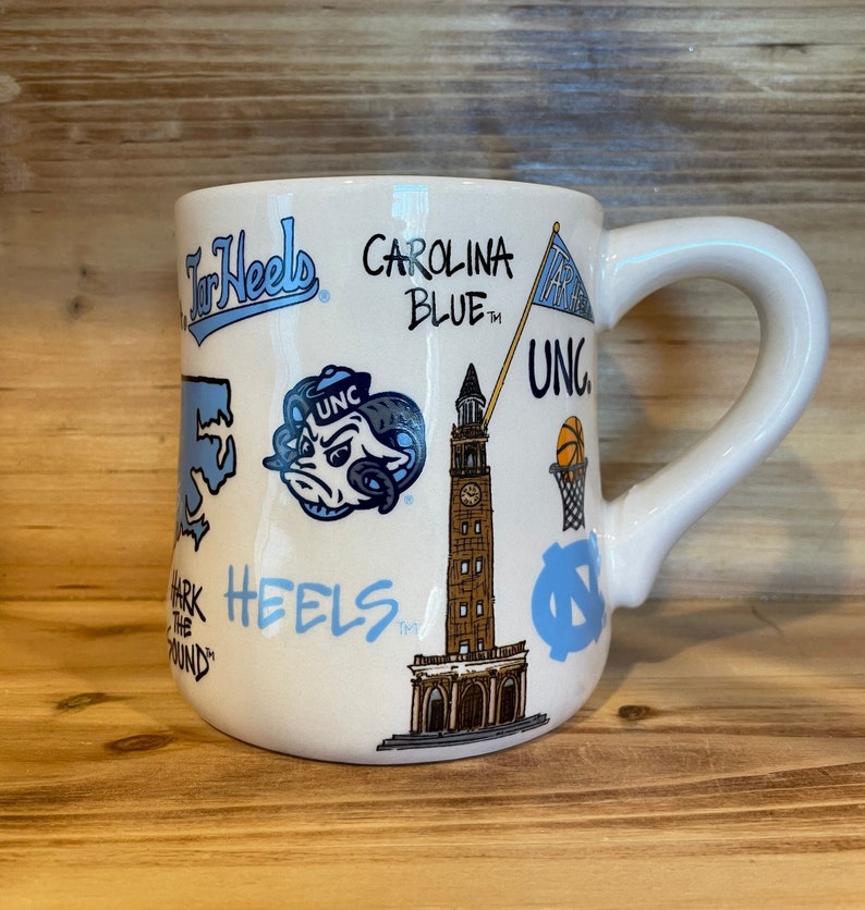 Peut inclure: Mug en c&eacute;ramique blanche orn&eacute; de graphiques et de texte bleu Carolina, dont "Tar Heels", "Carolina Blue" et "UNC". La tasse a une anse incurv&eacute;e et est d&eacute;cor&eacute;e d'illustrations d'un b&eacute;lier, d'un ballon de basket et d'une tour de l'horloge.