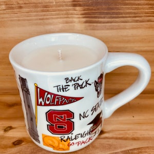 May include: A white ceramic mug candle with a wick. The mug is decorated with red and white graphics and text, including "WOLFPACK", "NC State", and "RALEIGH GO PACK". The candle is filled with a light-coloured wax.
