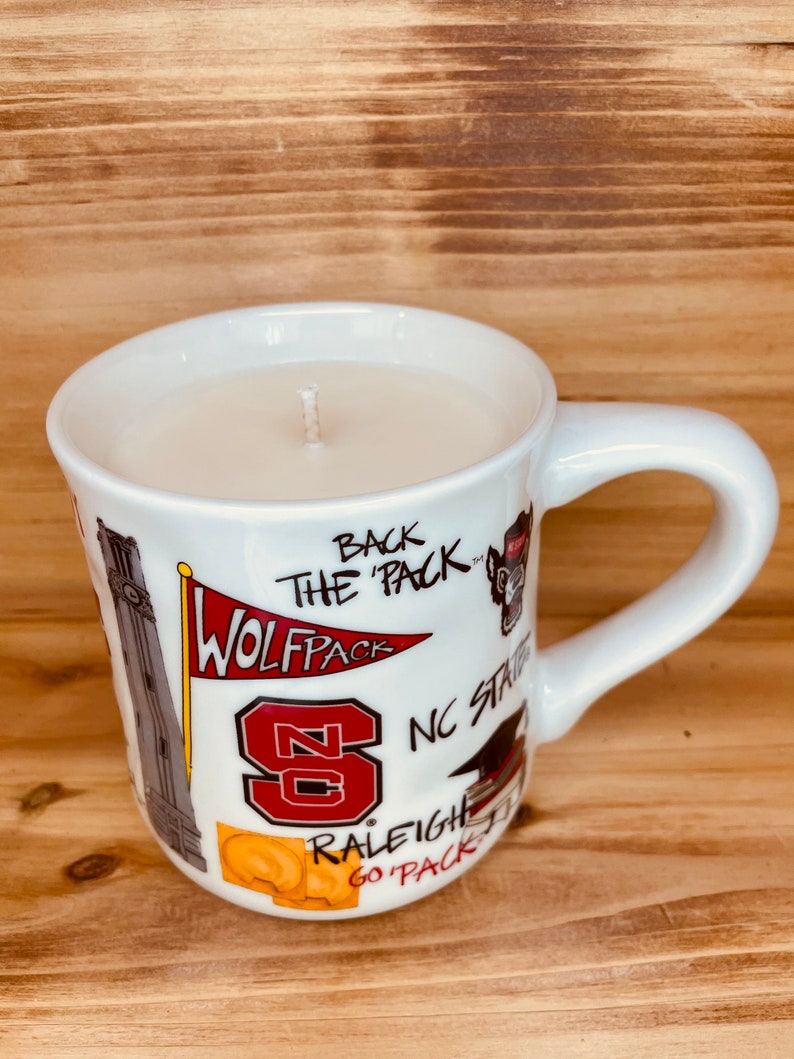 May include: A white ceramic mug candle with a wick. The mug is decorated with red and white graphics and text, including "WOLFPACK", "NC State", and "RALEIGH GO PACK". The candle is filled with a light-coloured wax.