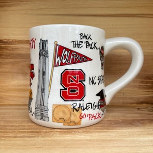 May include: White ceramic mug with a curved handle, decorated with illustrations and text related to NC State. Features a red and white Wolfpack flag, the letters "NC", and the words "Back the Pack".