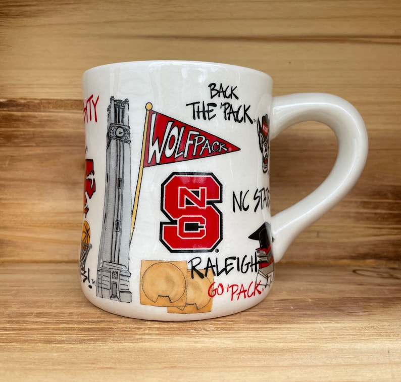 May include: White ceramic mug with a curved handle, decorated with illustrations and text related to NC State. Features a red and white Wolfpack flag, the letters "NC", and the words "Back the Pack".