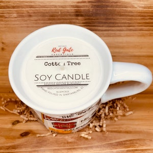 May include: A white mug candle with the text "Cotton Tree Soy Candle" and "Red Gate Essentials". The candle is hand-poured in Spartanburg, SC, and weighs 453g. The mug has a handle and is resting on brown packing material.