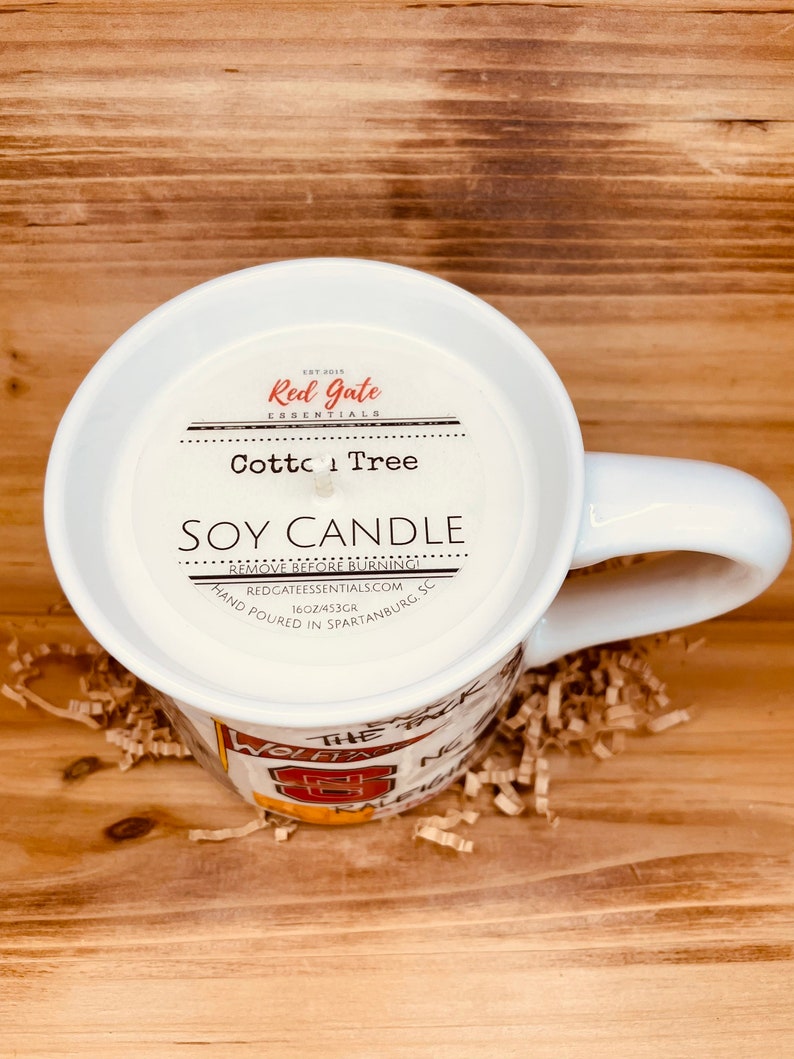 May include: A white mug candle with the text "Cotton Tree Soy Candle" and "Red Gate Essentials". The candle is hand-poured in Spartanburg, SC, and weighs 453g. The mug has a handle and is resting on brown packing material.