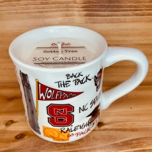 May include: A white ceramic mug filled with a soy candle. The mug has a handle and is decorated with the text "WOLFPACK", "NC STATE", and other graphics. The candle is labelled "Cotton Tree" and "SOY CANDLE".