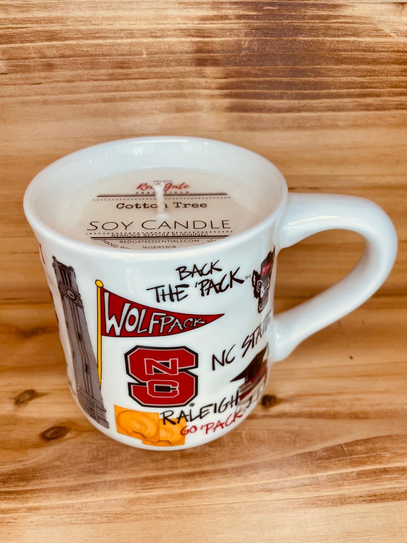 May include: A white ceramic mug filled with a soy candle. The mug has a handle and is decorated with the text "WOLFPACK", "NC STATE", and other graphics. The candle is labelled "Cotton Tree" and "SOY CANDLE".