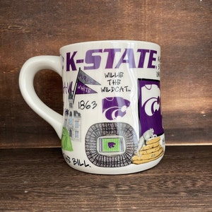 May include: White ceramic coffee mug with a purple and white design featuring the text "K-STATE" and various images related to Kansas State University, including the school mascot, Willie the Wildcat, the university's football stadium, and the school's founding date, 1863.