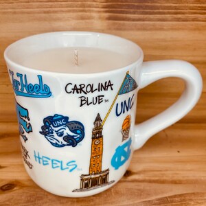 Peut inclure: Bougie dans une tasse en c&eacute;ramique blanche avec une anse. La tasse est orn&eacute;e de logos bleu Carolina, des inscriptions &laquo; Tar Heels &raquo; et &laquo; Carolina Blue &raquo;, ainsi que d'autres graphiques. La bougie repose sur une surface en bois.