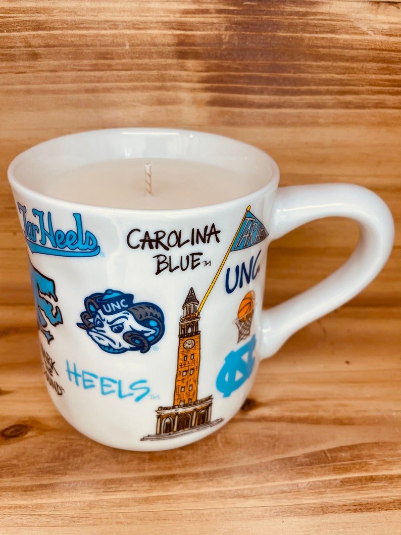 Peut inclure: Bougie dans une tasse en c&eacute;ramique blanche avec une anse. La tasse est orn&eacute;e de logos bleu Carolina, des inscriptions &laquo; Tar Heels &raquo; et &laquo; Carolina Blue &raquo;, ainsi que d'autres graphiques. La bougie repose sur une surface en bois.