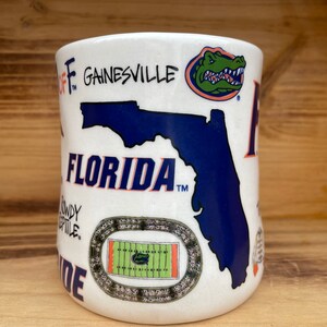 May include: White ceramic mug with a blue and orange Florida Gators design. The mug features the state of Florida, a gator mascot, a football field, and the words "Florida" and "Gainesville".
