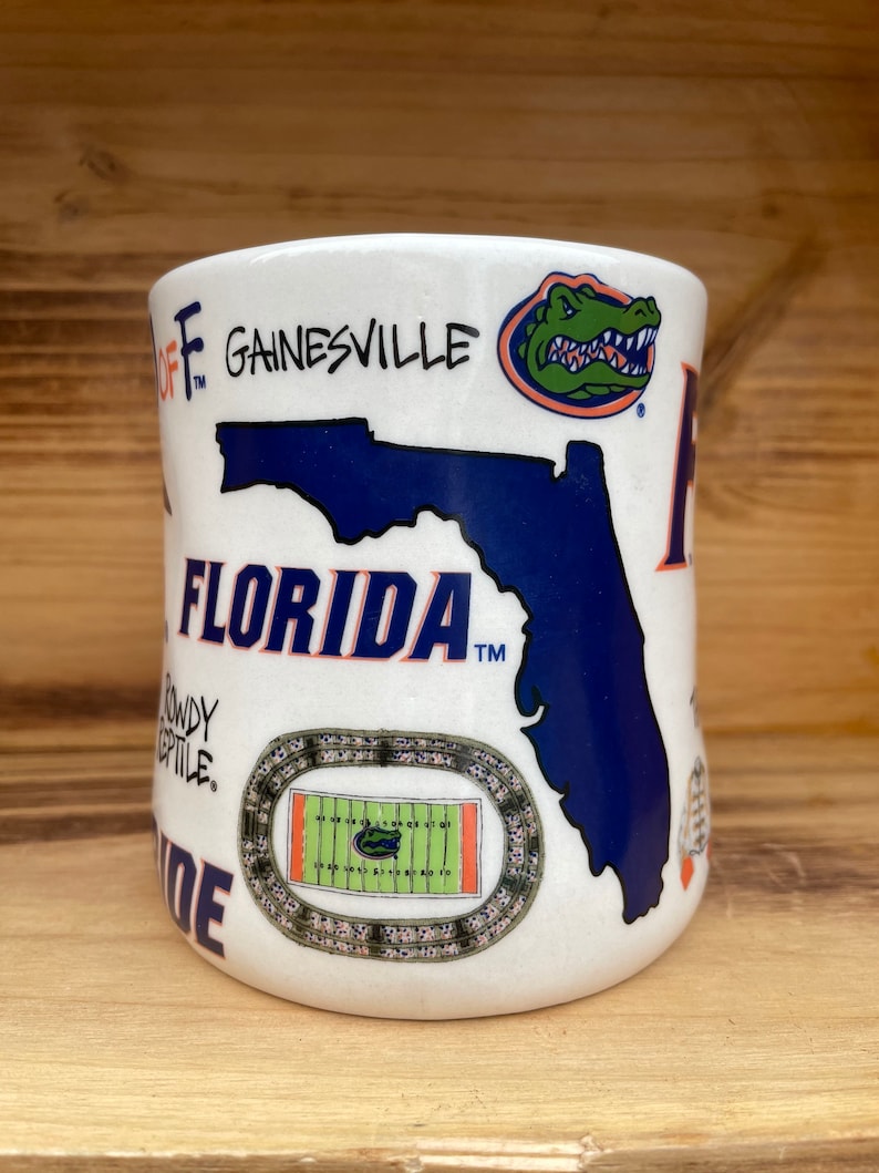 May include: White ceramic mug with a blue and orange Florida Gators design. The mug features the state of Florida, a gator mascot, a football field, and the words "Florida" and "Gainesville".
