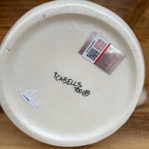 May include: White ceramic mug with the text "TCABELLS Too&reg;" and a small sticker with a crown design on the bottom. The mug also has a sticker with a barcode and the text "A654580369".