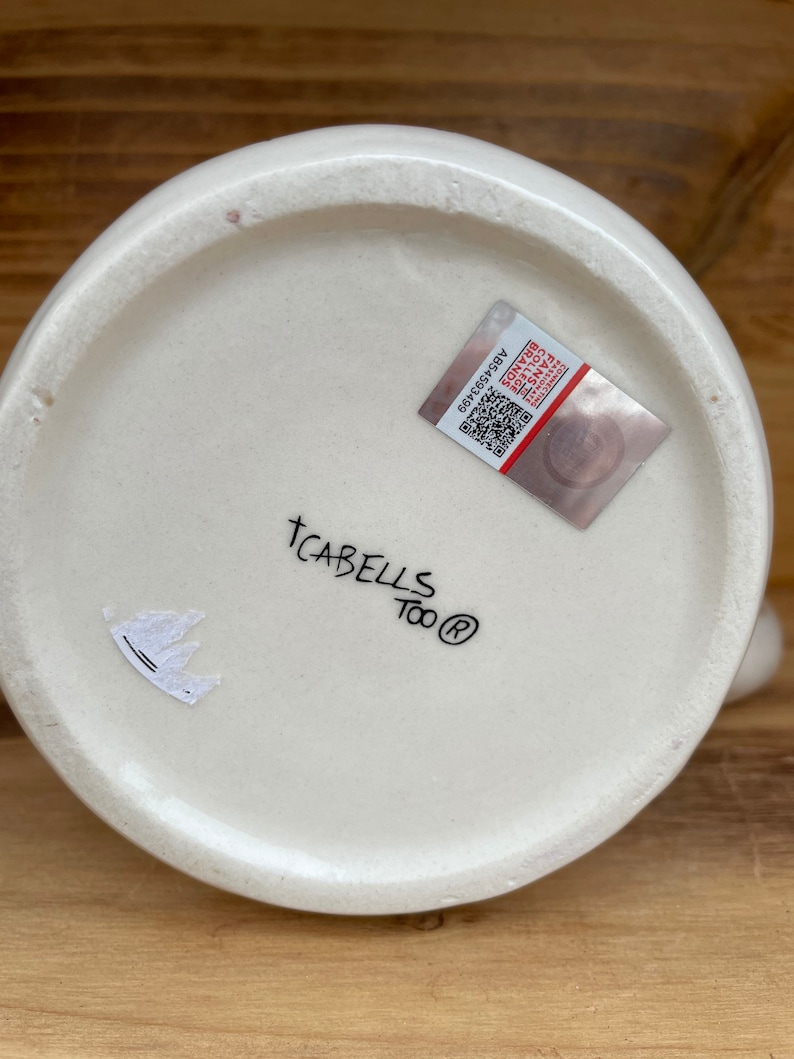 May include: White ceramic mug with the text "TCABELLS Too&reg;" and a small sticker with a crown design on the bottom. The mug also has a sticker with a barcode and the text "A654580369".