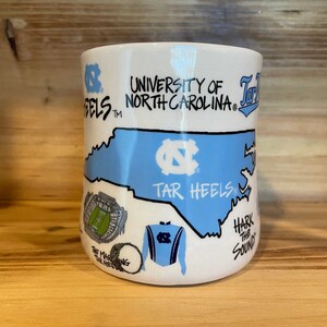 Peut inclure: Mug en c&eacute;ramique blanche avec un contour bleu clair de la Caroline du Nord, les mots "University of North Carolina" et "Tar Heels". La tasse comprend des illustrations d'un stade, d'un tambour et d'un uniforme de fanfare.