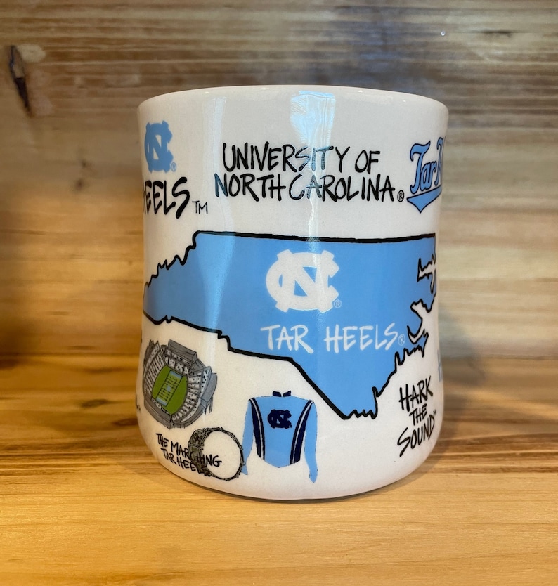 Peut inclure: Mug en c&eacute;ramique blanche avec un contour bleu clair de la Caroline du Nord, les mots "University of North Carolina" et "Tar Heels". La tasse comprend des illustrations d'un stade, d'un tambour et d'un uniforme de fanfare.